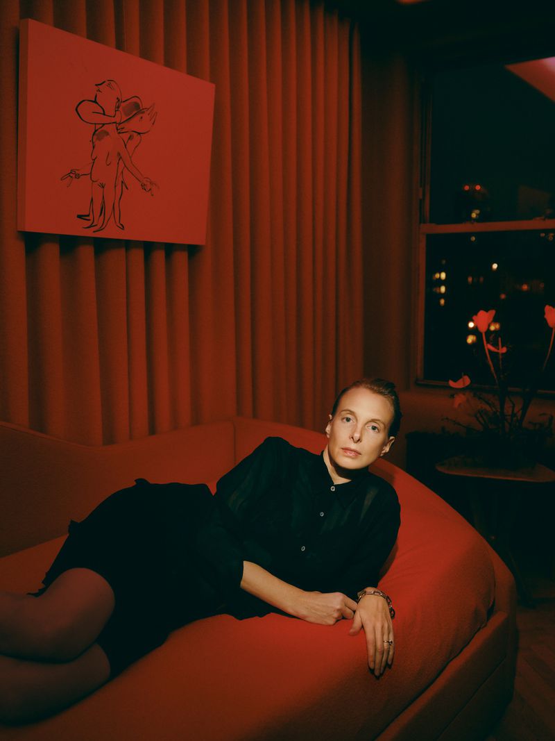 Adult female reclines on a chaise lounge in a red-toned interior with a modern artwork and city lights visible through a window at night.