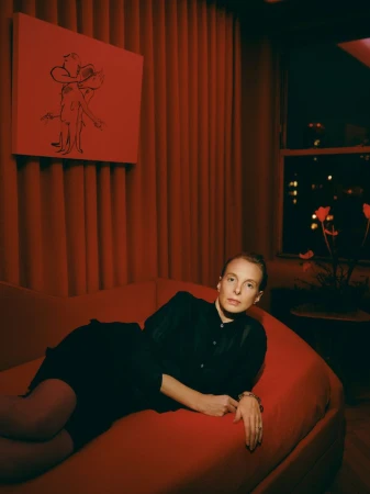 Adult female reclines on a chaise lounge in a red-toned interior with a modern artwork and city lights visible through a window at night.