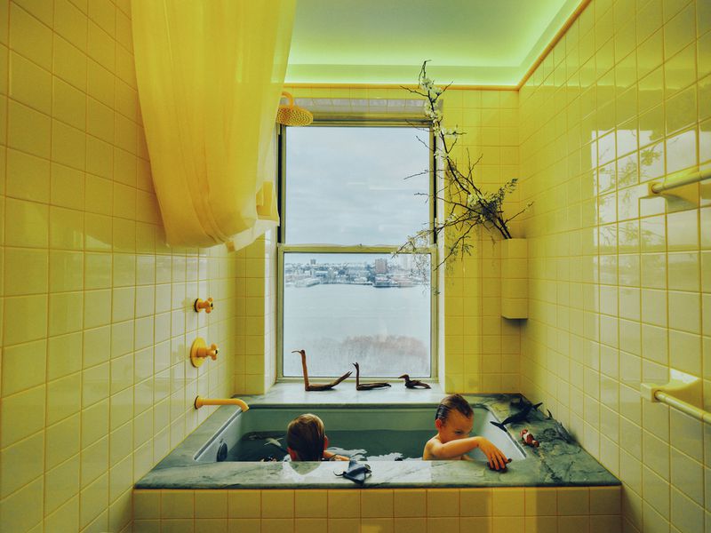Two kids enjoy bathtime surrounded by yellow tile, with toy animals and a river view visible through a window. Interior, natural light.