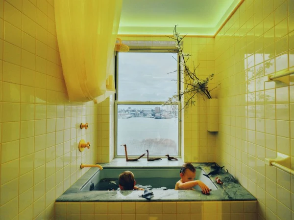 Two kids enjoy bathtime surrounded by yellow tile, with toy animals and a river view visible through a window. Interior, natural light.