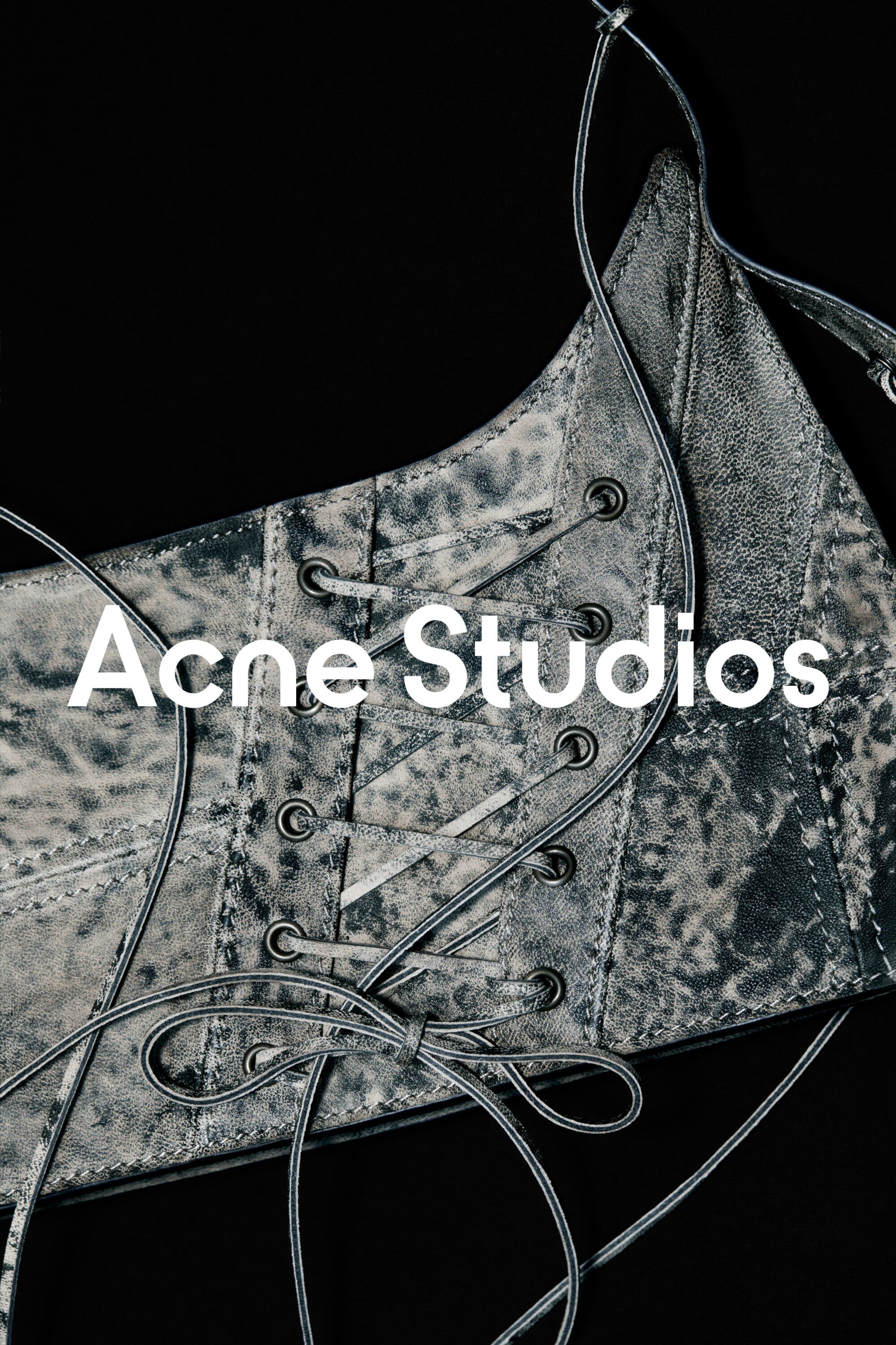 Close-up of a silver corset with black lacing against a dark backdrop, featuring the 'Acne Studios' logo, showcasing fashion and design.
