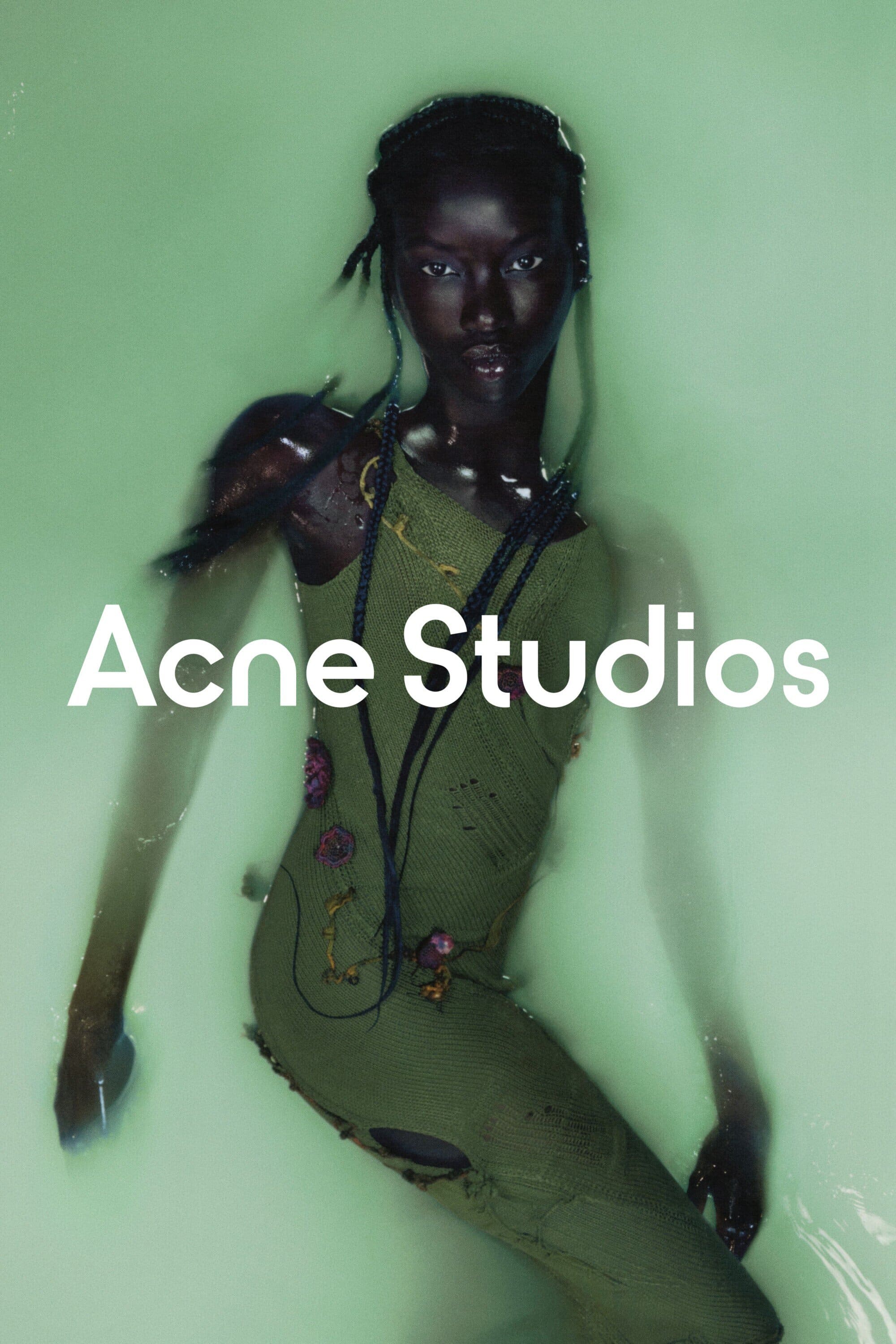 A model poses in an indoor studio against a pastel green backdrop, wearing an olive green dress with water swirling around her.