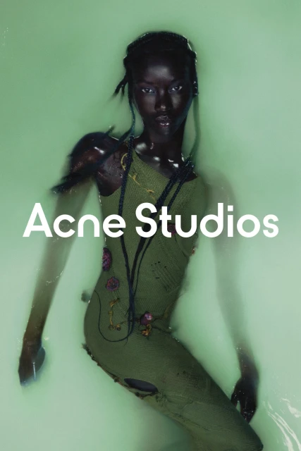 A model poses in an indoor studio against a pastel green backdrop, wearing an olive green dress with water swirling around her.