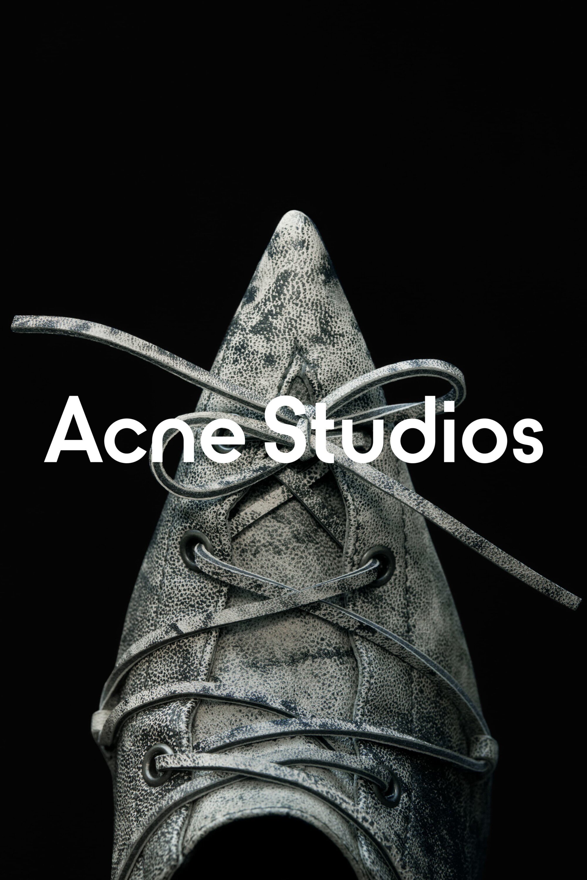 Close-up studio shot of a gray leather shoe with laces, set against a stark black backdrop, with the 'Acne Studios' logo overlaid.