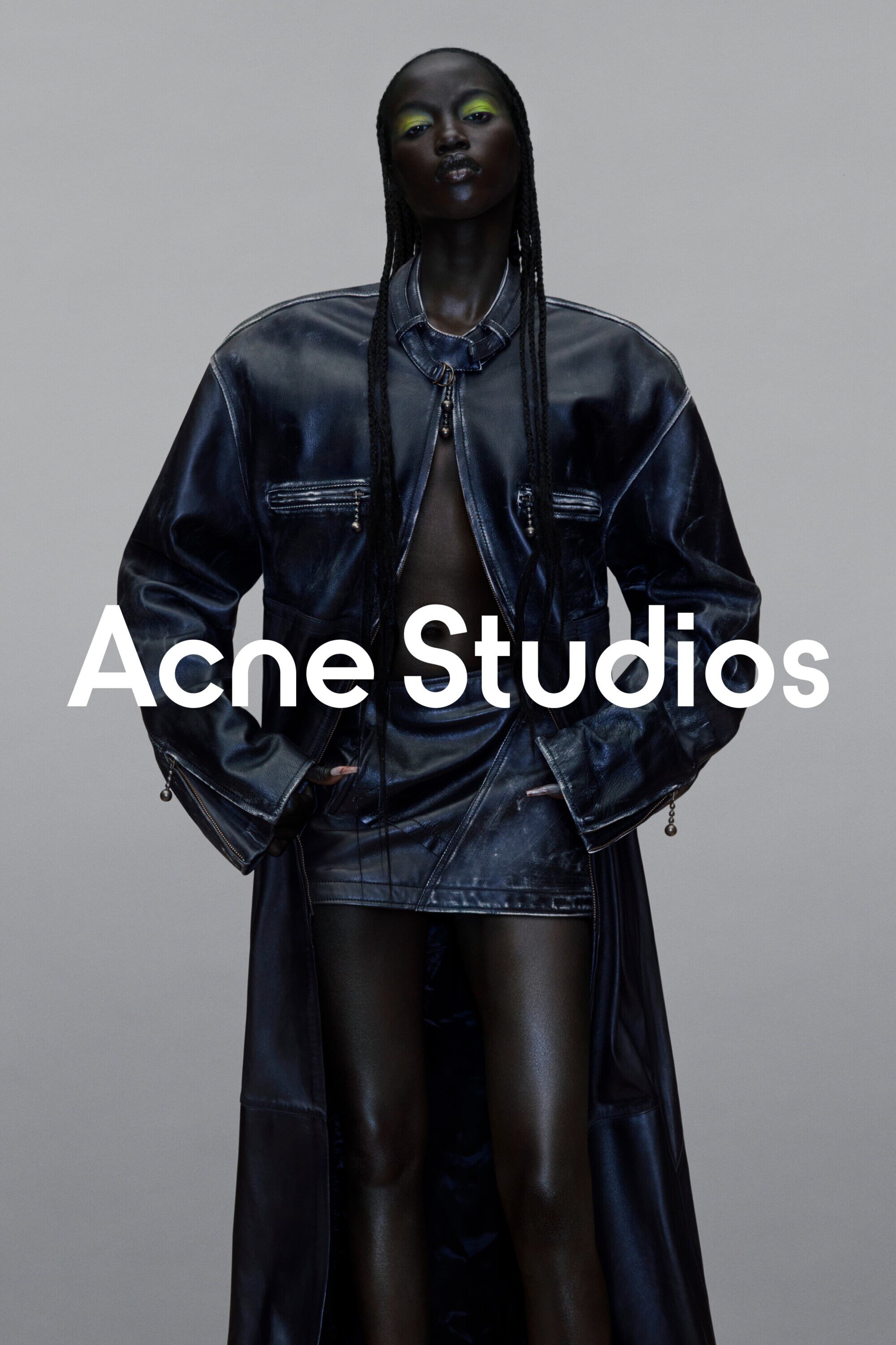 Dark-skinned fashion model poses in a shiny blue coat and mini skirt with green eyeshadow against a neutral grey background.