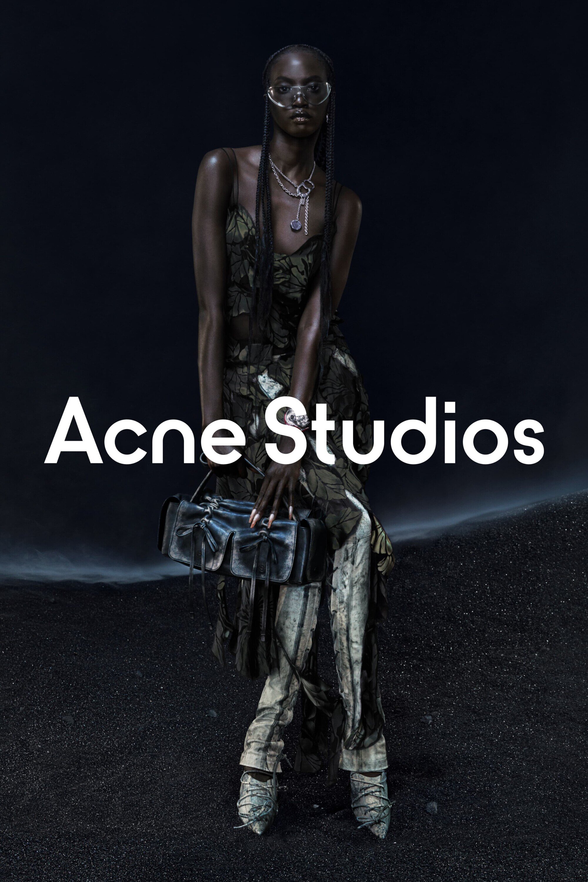 A dark-skinned model poses against a dark backdrop, wearing a dress, boots, and jewelry while holding a handbag for Acne Studios.