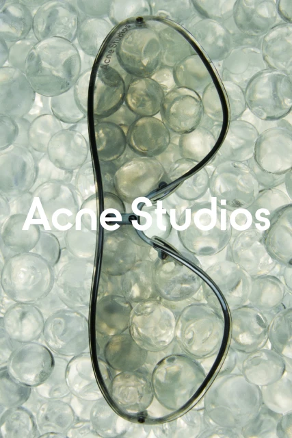 Sunglasses float above a collection of clear glass beads, accented by the 'Acne Studios' logo, which is featured in white text.