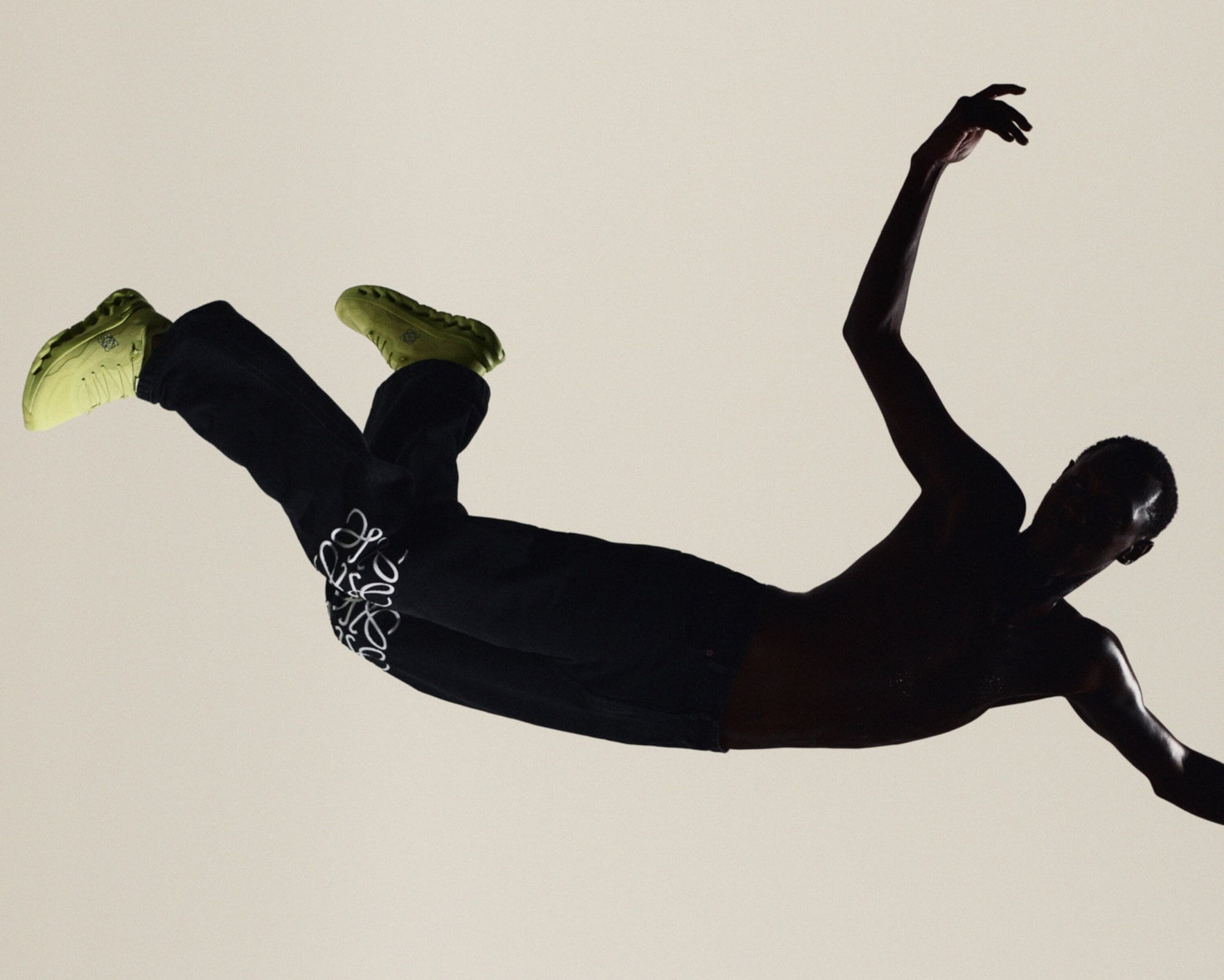 Shirtless male dancer leaping horizontally in mid-air wearing black Loewe sweatpants and lime green sneakers against a beige background.