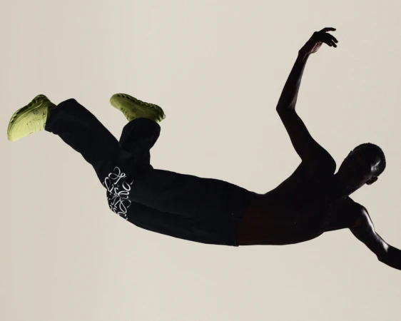 Shirtless male dancer leaping horizontally in mid-air wearing black Loewe sweatpants and lime green sneakers against a beige background.