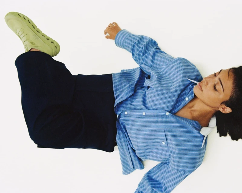 Woman in blue striped shirt and black pants lies on a white surface, wearing green shoes. Studio fashion portrait with natural light.