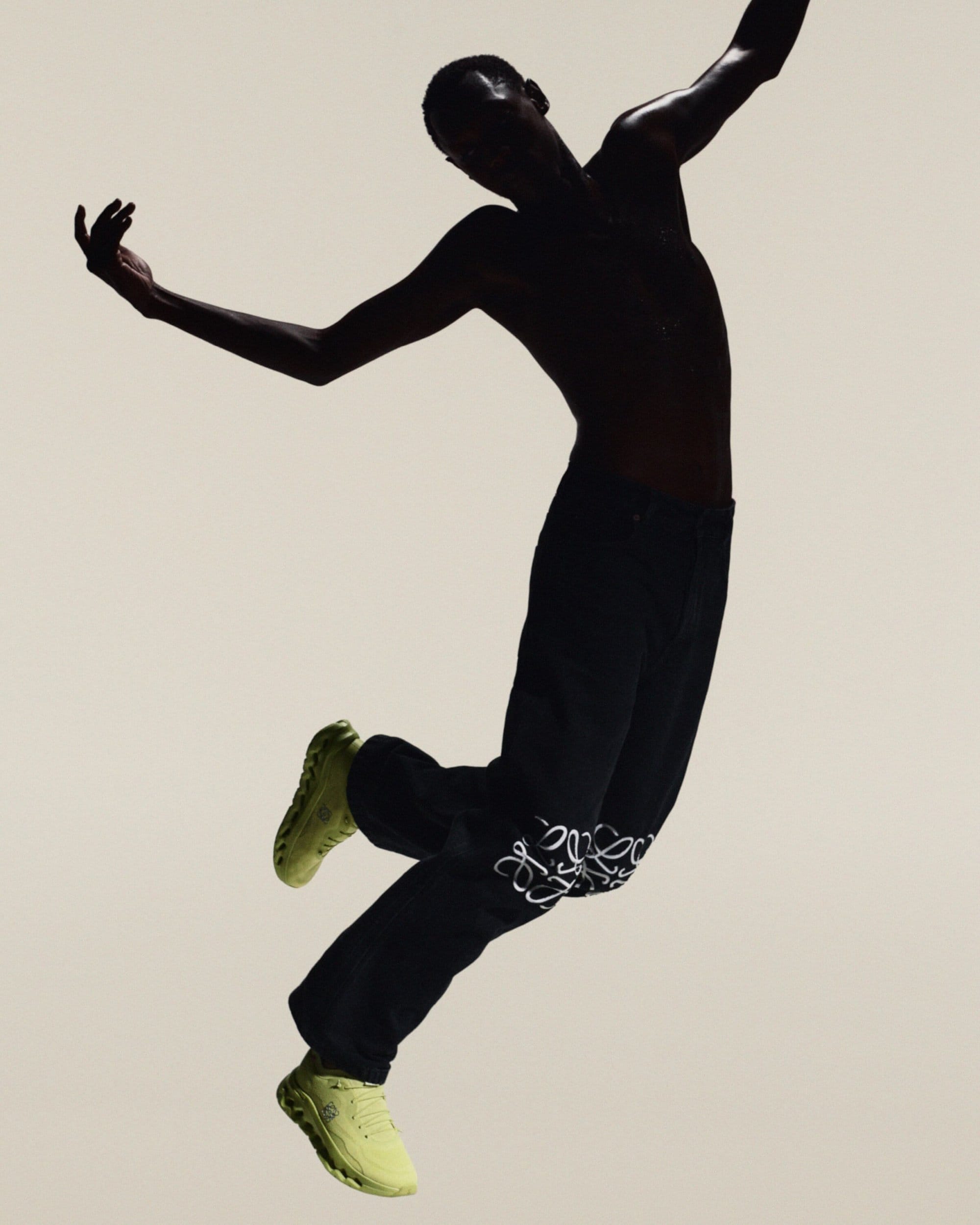 Adult male in mid-air, wearing dark pants with white embroidery, light-colored sneakers, set against a neutral background in a studio shot.