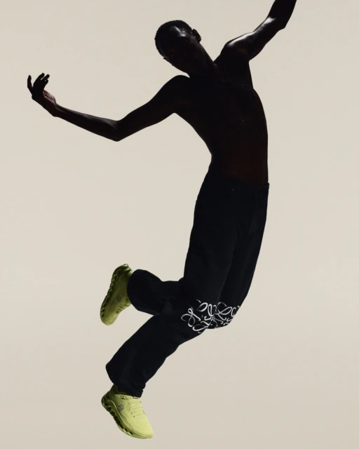 Adult male in mid-air, wearing dark pants with white embroidery, light-colored sneakers, set against a neutral background in a studio shot.