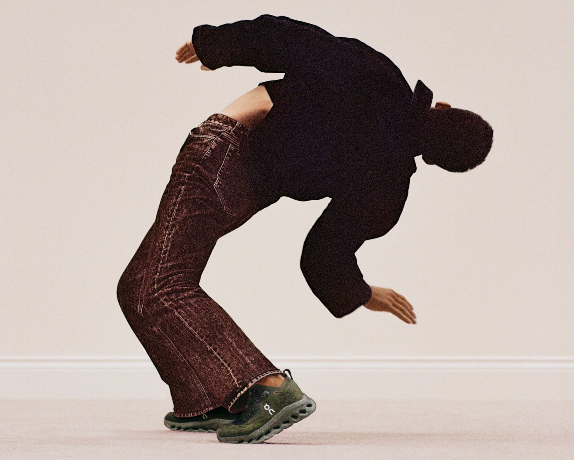 Man in dark clothing and jeans performing a backbend, wearing olive green running shoes against a neutral-toned background.