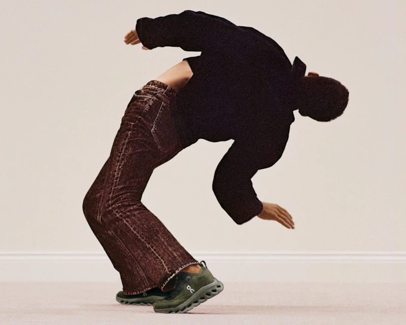 Man in dark clothing and jeans performing a backbend, wearing olive green running shoes against a neutral-toned background.