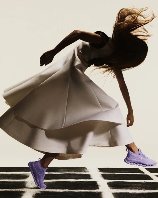 Woman in motion wearing a white dress and lavender sneakers on a black and white geometric surface against an off-white background.