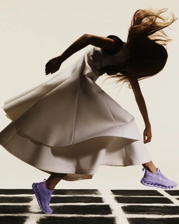 Woman in motion wearing a white dress and lavender sneakers on a black and white geometric surface against an off-white background.
