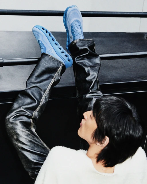 Person wearing black leather-look pants and light blue ON running shoes in studio setting with black bench and minimalist aesthetic.