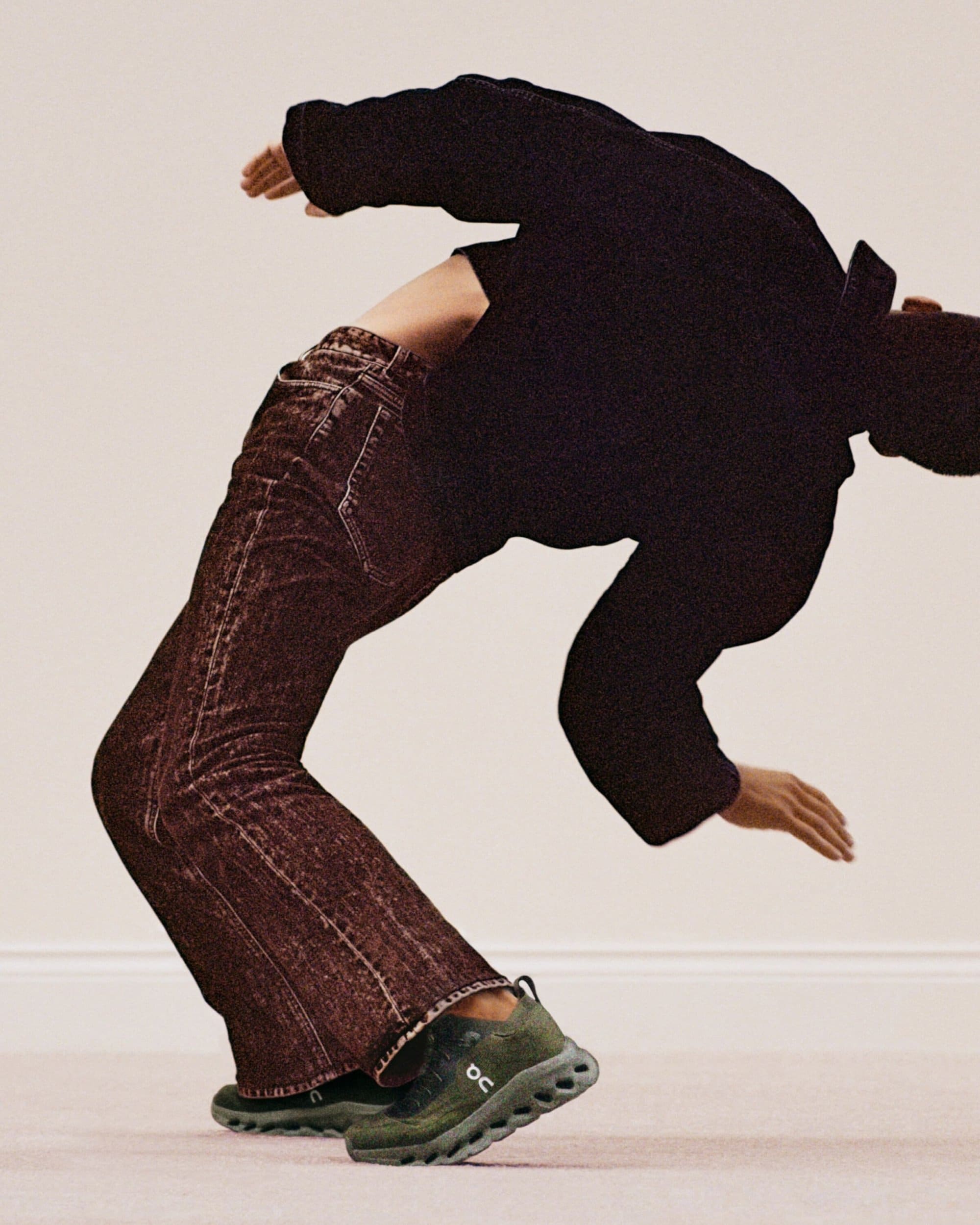 Person in a backbend, wearing a black shirt, brown jeans, and green sneakers against a neutral background with soft lighting.