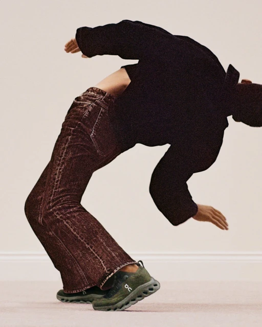 Person in a backbend, wearing a black shirt, brown jeans, and green sneakers against a neutral background with soft lighting.