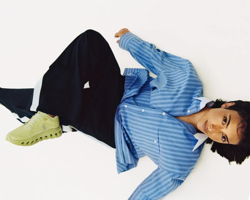 Model poses with a chartreuse running shoe, wearing a blue striped shirt and black pants on a white surface, showcasing fashion apparel and footwear.