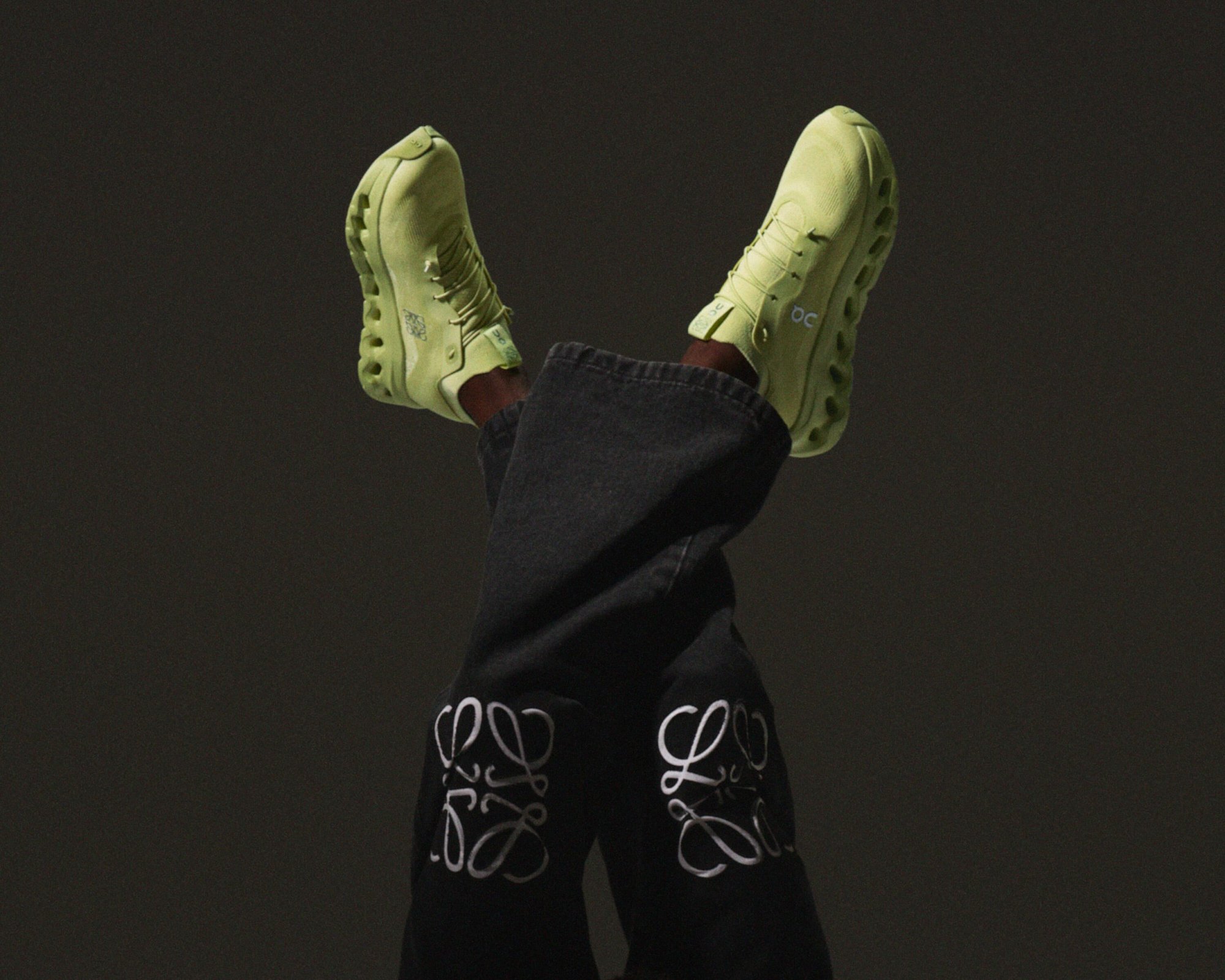 Elevated shot of person wearing green On Running sneakers and designer jeans with white logo on a grey studio background.