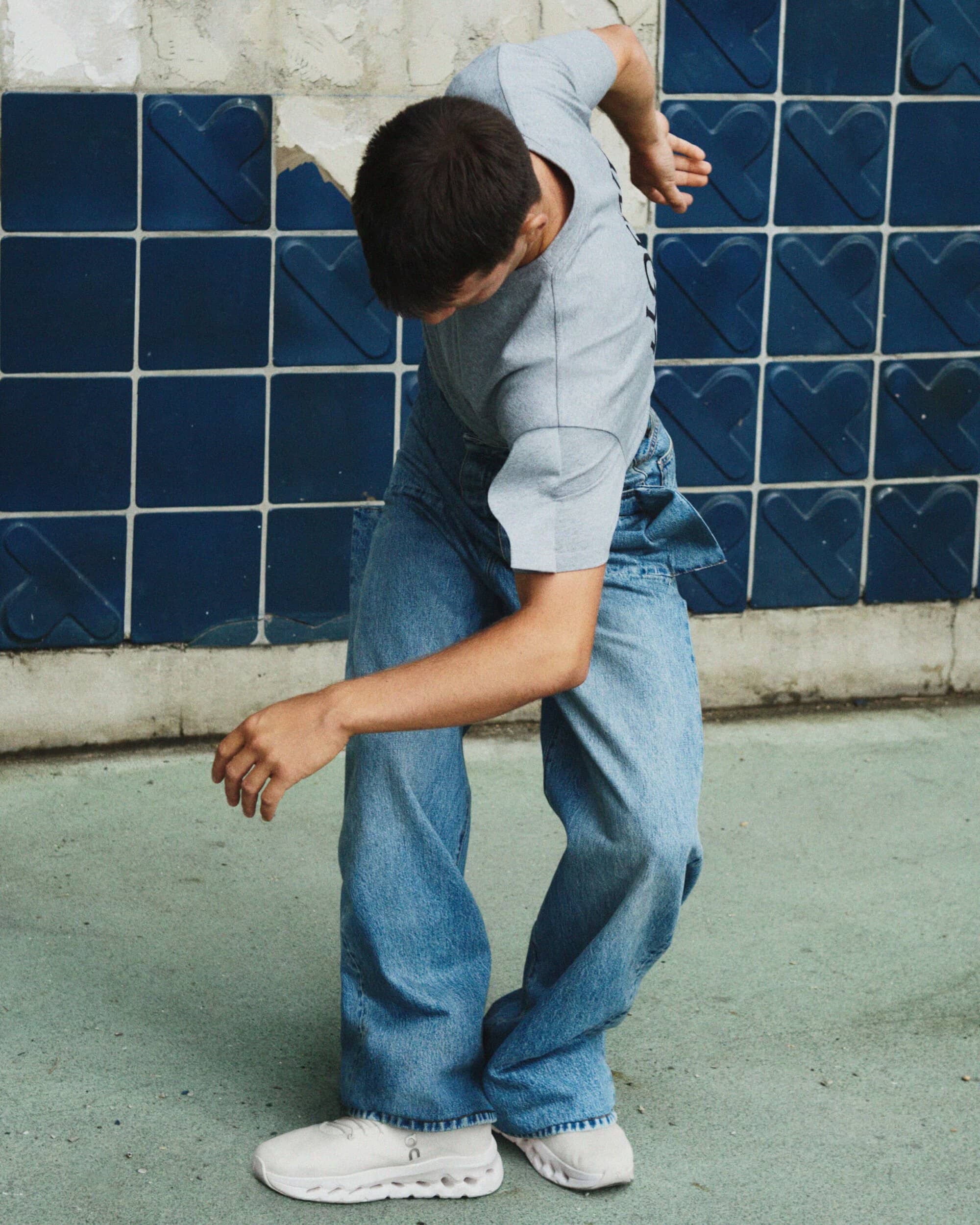 Young adult dancing in blue jeans and grey t-shirt against a tiled wall, showcasing contemporary movement and style.