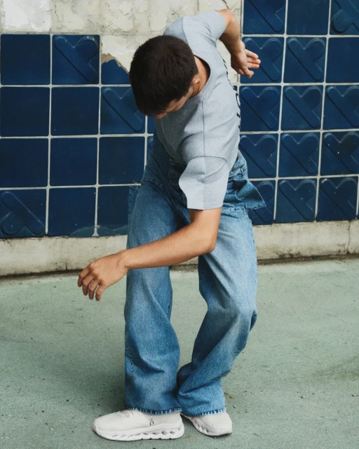 Young adult dancing in blue jeans and grey t-shirt against a tiled wall, showcasing contemporary movement and style.