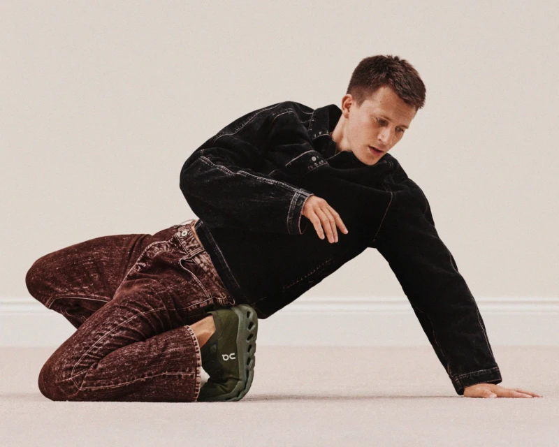 An adult male is lying on the floor, wearing a black denim jacket, dark red jeans, and green running shoes, against a light beige background.