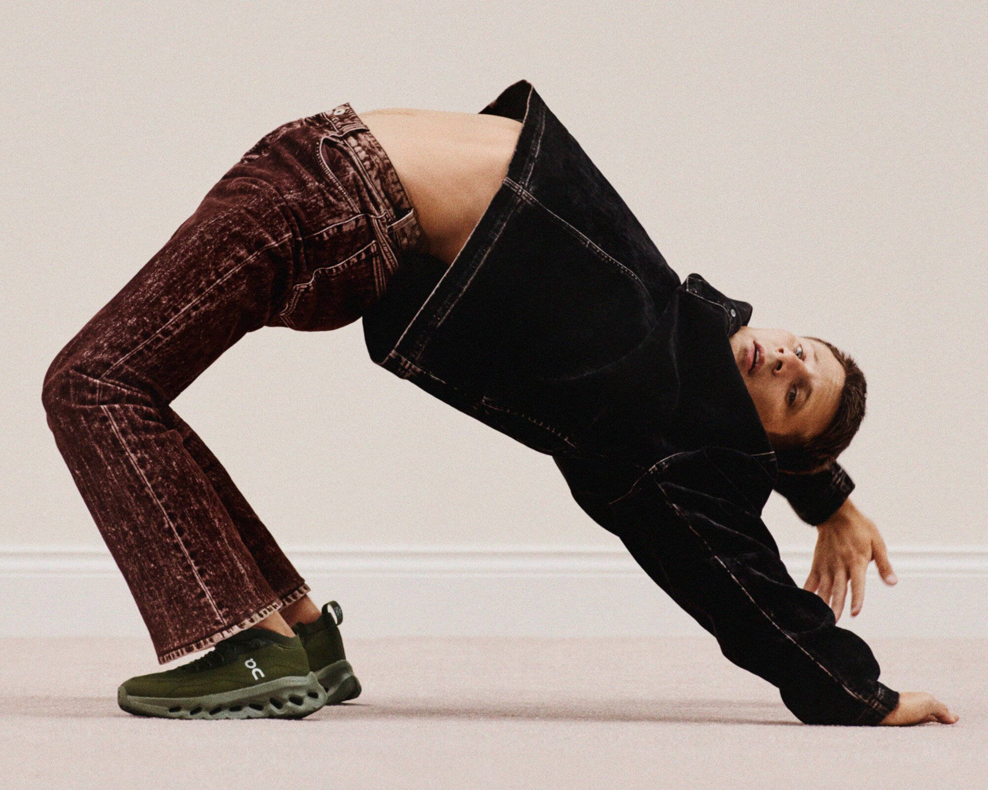 Man in bridge pose wearing black jacket, burgundy jeans and green running shoes on cream carpet in a modern, minimalist interior.
