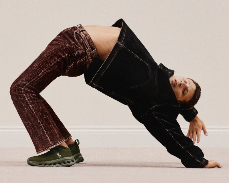Man in bridge pose wearing black jacket, burgundy jeans and green running shoes on cream carpet in a modern, minimalist interior.