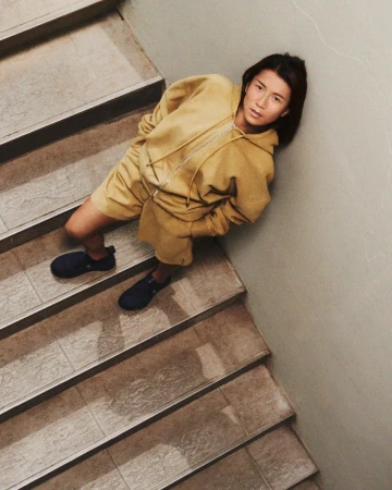 Adult woman wearing yellow sweatsuit and dark sneakers poses on a tiled staircase with a light gray wall in the background.