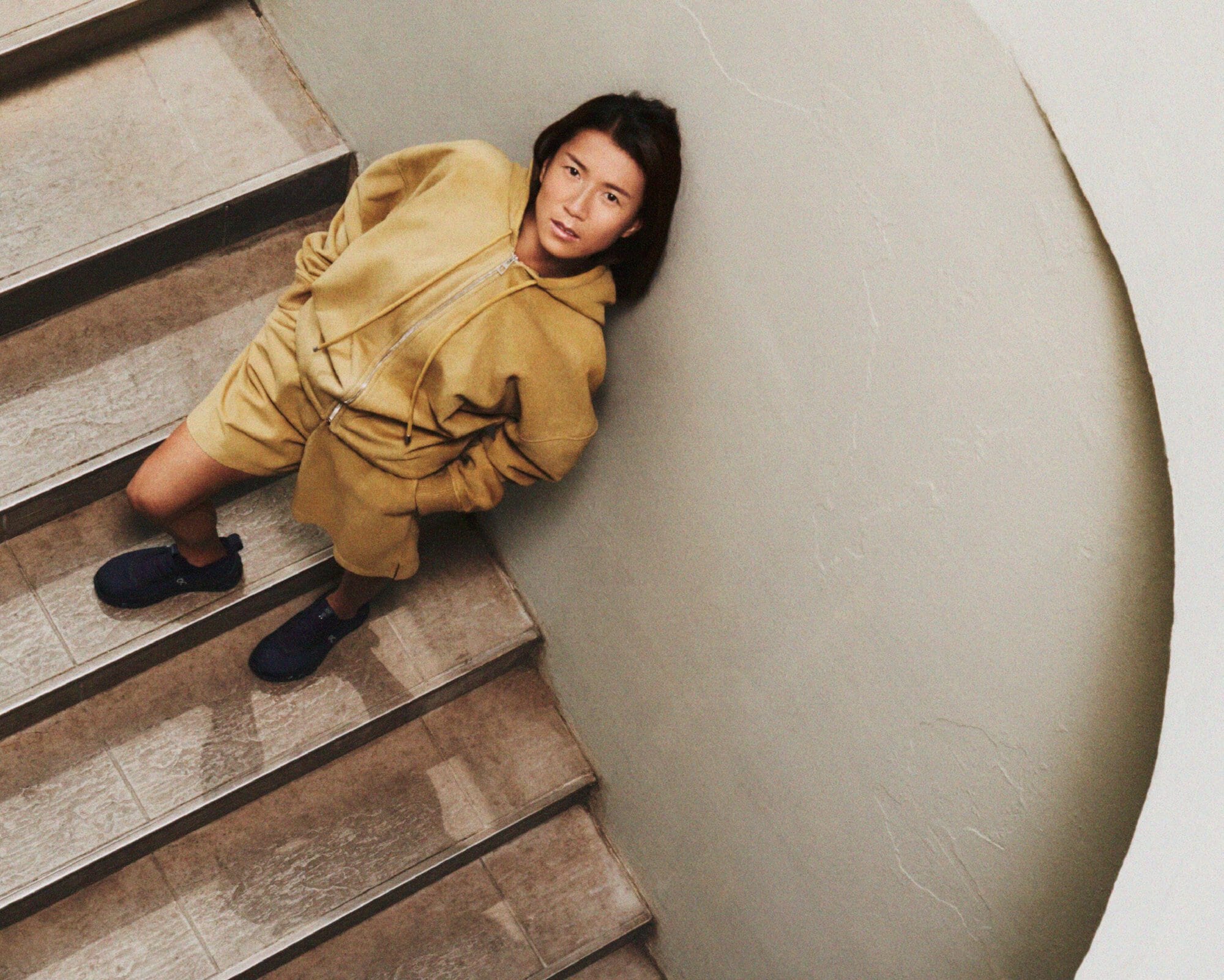 Person in yellow hooded sweatshirt and matching shorts standing on stairs, wearing dark sneakers, captured from a high-angle perspective.