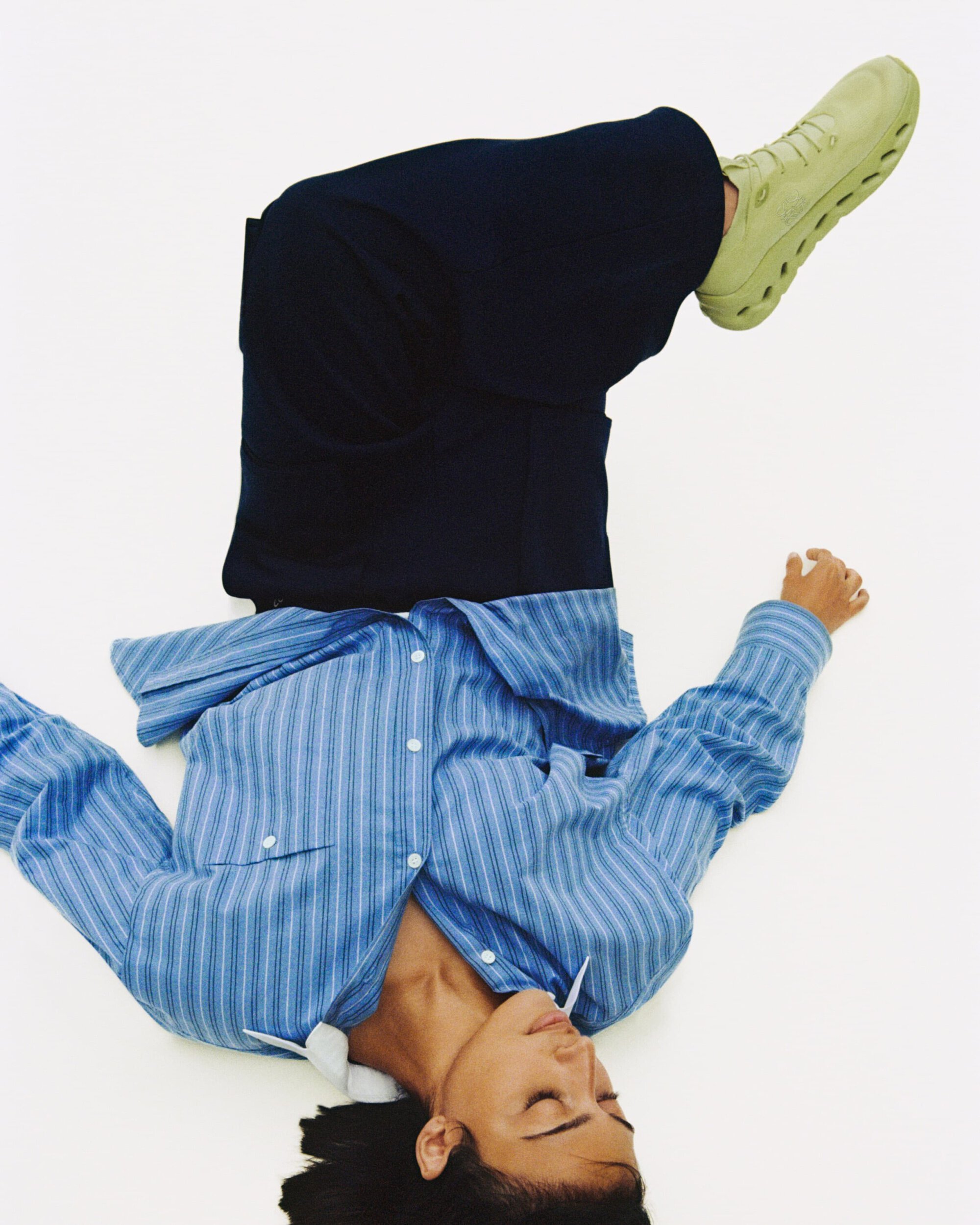 Model lying on back against a white background wearing a blue striped shirt, dark trousers, and pale green sneakers