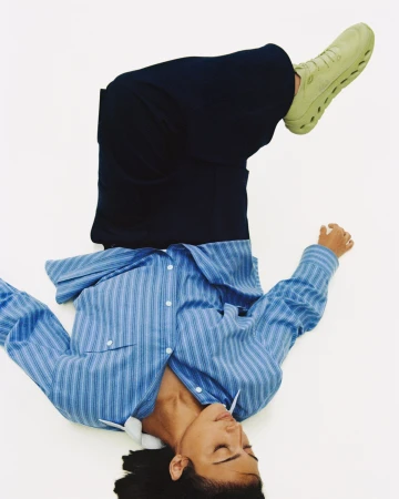 Model lying on back against a white background wearing a blue striped shirt, dark trousers, and pale green sneakers