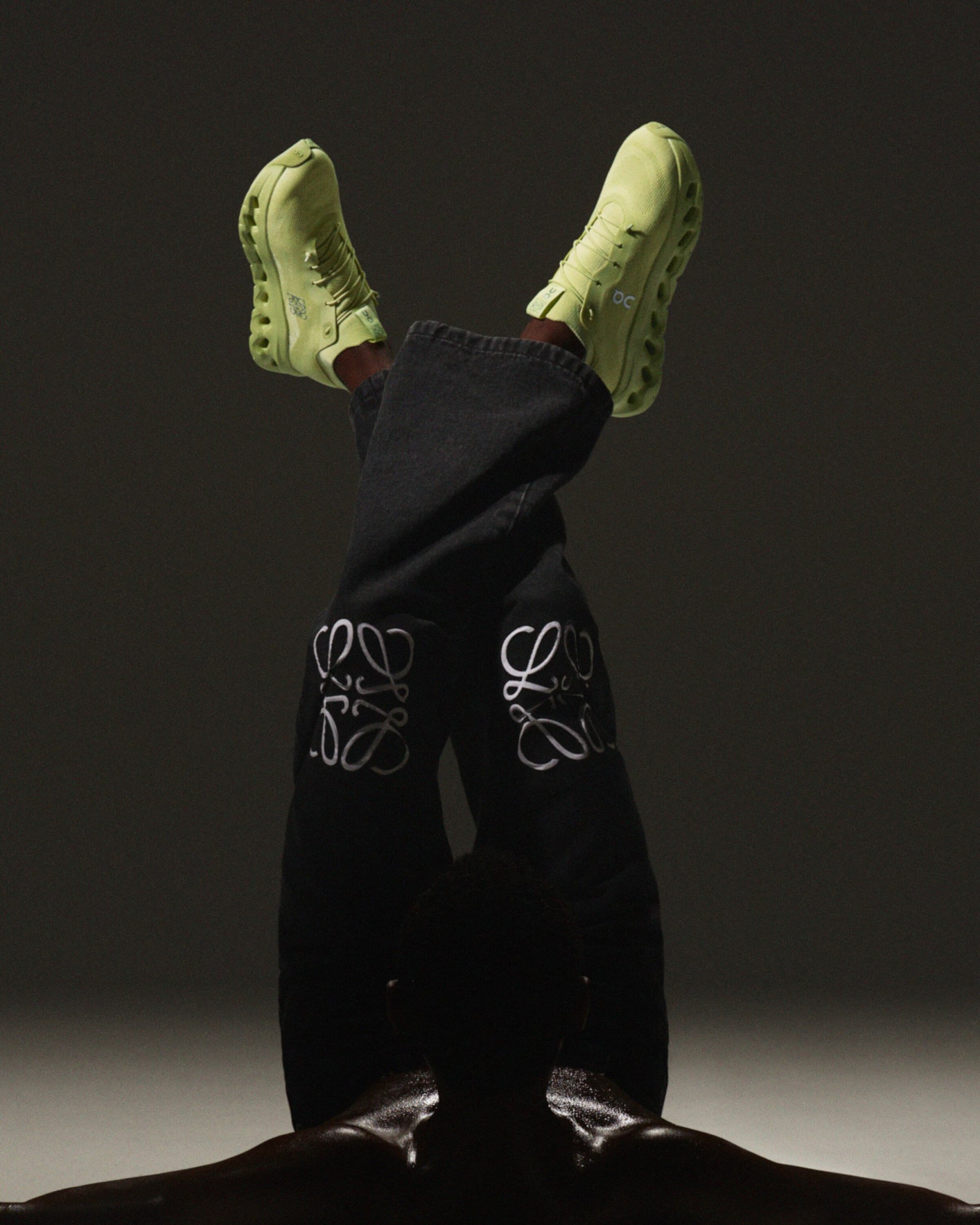 Upside-down legs wearing black denim jeans with white embroidered logos and light green sneakers against a dark studio background.