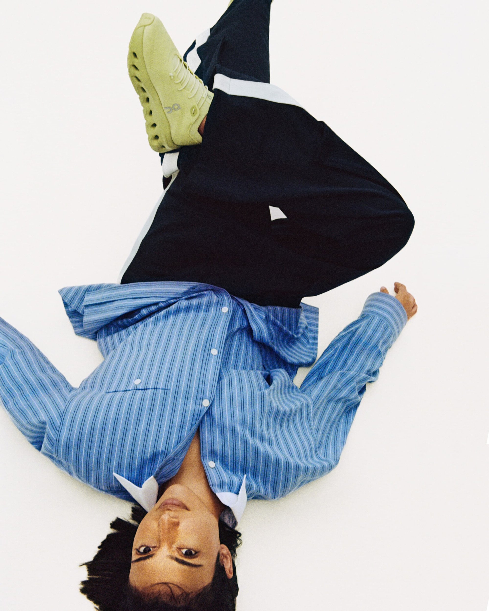 Upside-down portrait of a person lying on white background, wearing a blue striped shirt, black pants, and chartreuse sneakers.