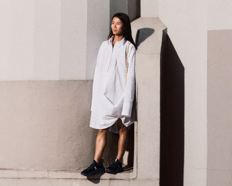 Woman in a white shirt dress poses outdoors in neutral light against a minimalist wall, showcasing a modern and relaxed fashion style.