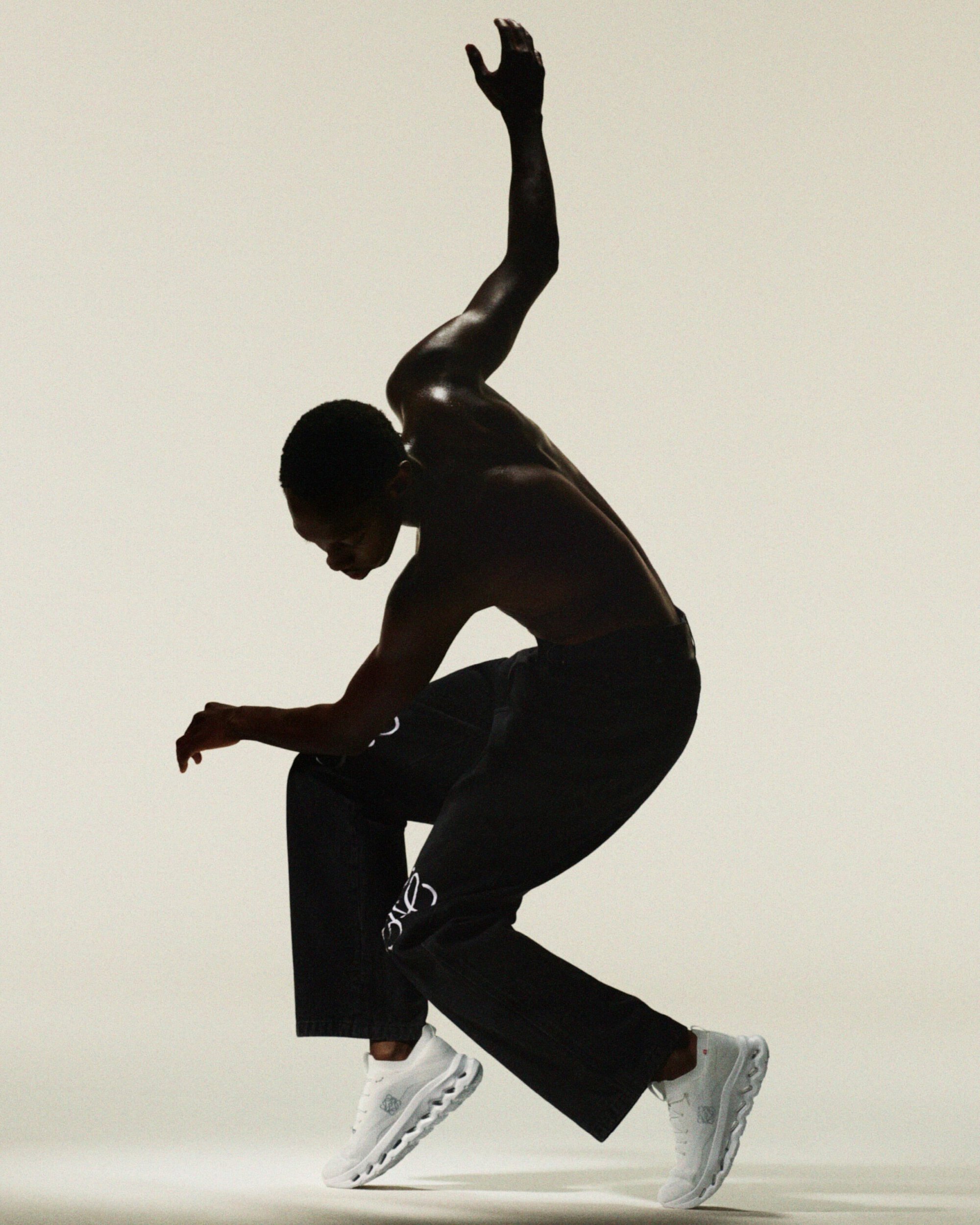 Athletic African-American male strikes dynamic pose in black pants and white sneakers against a light background, showcasing movement and style.