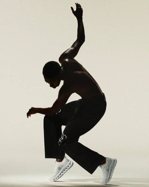 Athletic African-American male strikes dynamic pose in black pants and white sneakers against a light background, showcasing movement and style.