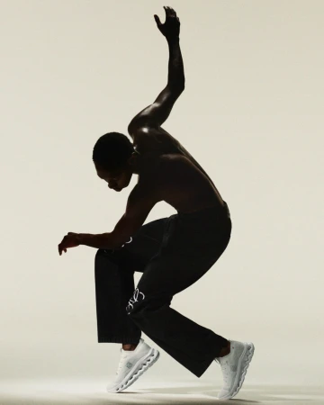 Athletic African-American male strikes dynamic pose in black pants and white sneakers against a light background, showcasing movement and style.