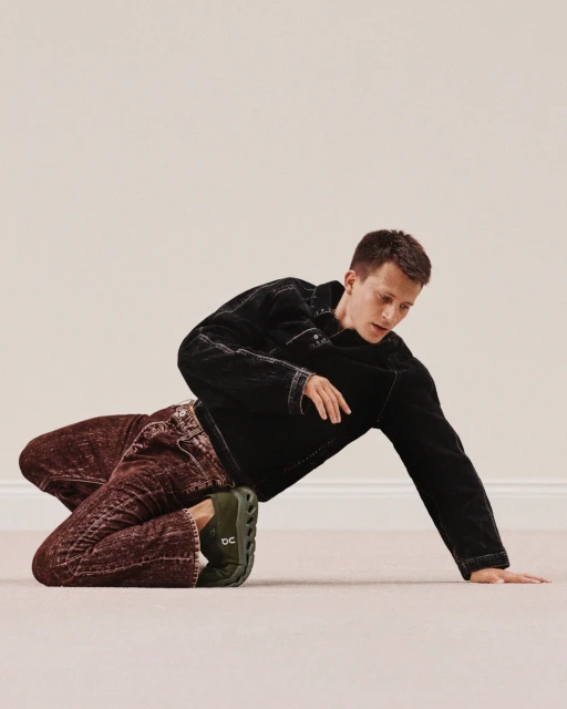 Athletic man in black denim jacket and dark jeans poses on the floor with a pair of dark green running shoes; neutral backdrop.