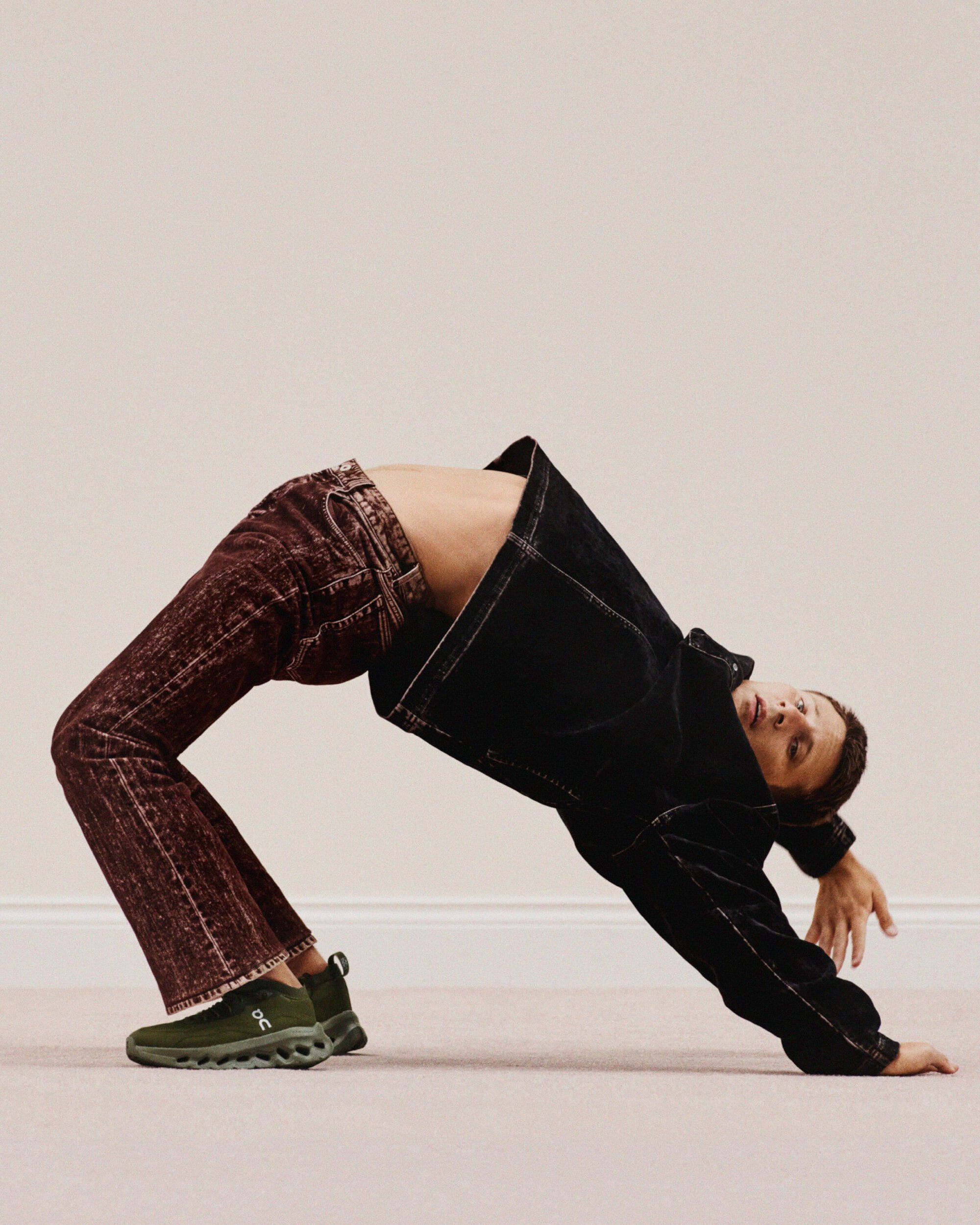 Adult performs a backbend wearing jeans, a black jacket, and green sneakers against a neutral wall and floor. One person in the image.