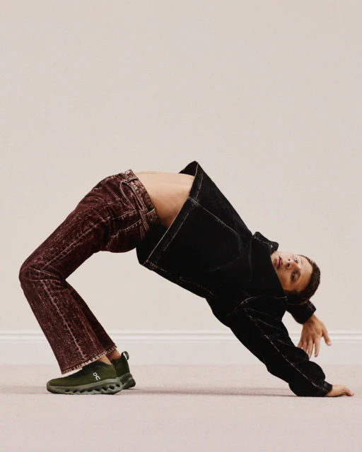 Adult performs a backbend wearing jeans, a black jacket, and green sneakers against a neutral wall and floor. One person in the image.