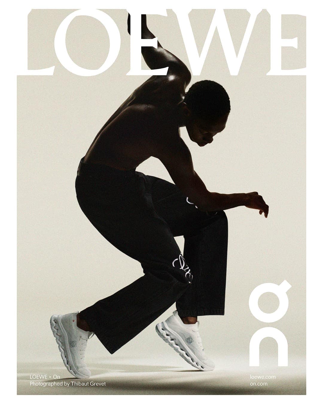 An athletic adult male model is posing indoors wearing black pants and white running shoes next to large brand logos.