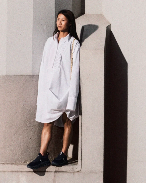 Adult woman in a white shirt dress and black running shoes poses against a concrete wall, creating a fashion-forward and sporty look.