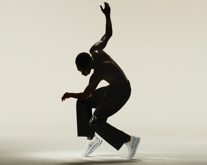 Shirtless man dynamically posing on tiptoe wearing black trousers and white sneakers against an off-white seamless background.