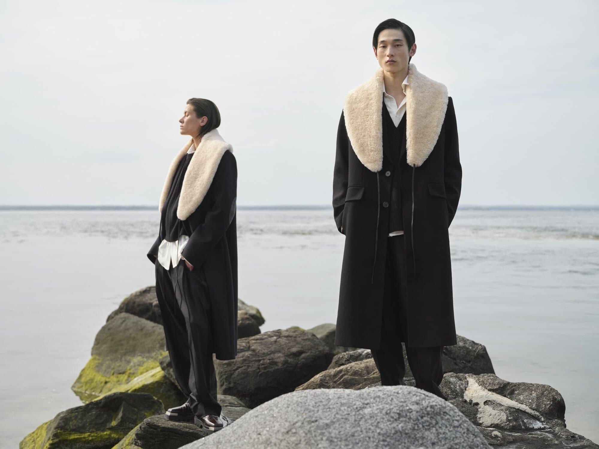 Two people in coats with fur collars stand on mossy rocks by the ocean under an overcast sky, showcasing fashion apparel.