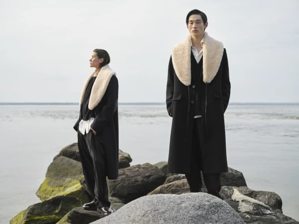 Two people in coats with fur collars stand on mossy rocks by the ocean under an overcast sky, showcasing fashion apparel.