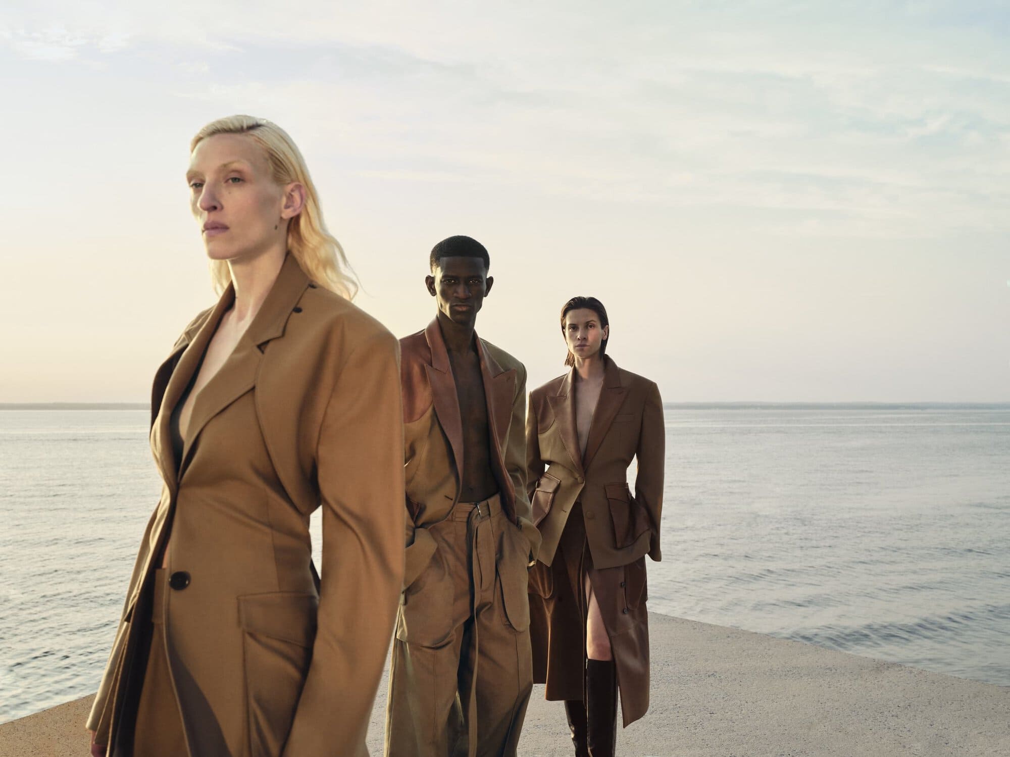 Three fashion models stand on a pier in tan outfits against a serene ocean backdrop under a bright sky, showcasing modern style.