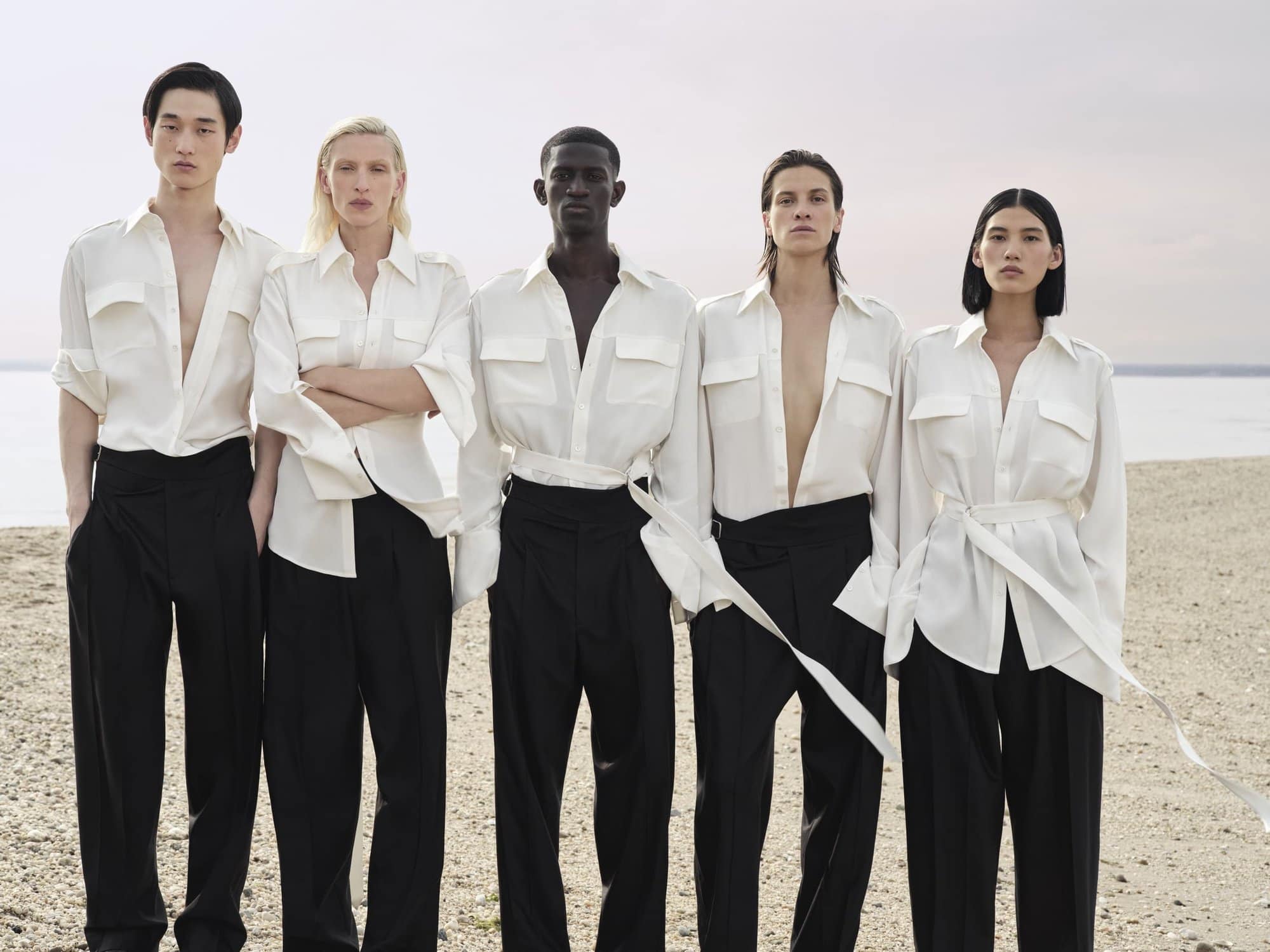 Fashion editorial featuring five models in white shirts and black pants posing on a beach with an overcast sky in the background.
